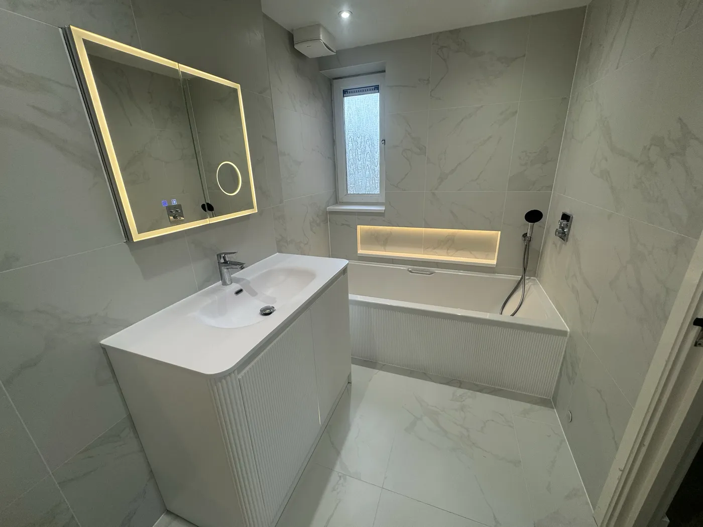 Marble-effect bathroom renovation with illuminated mirror, curved white vanity, fitted bath and LED-lit recessed niche.