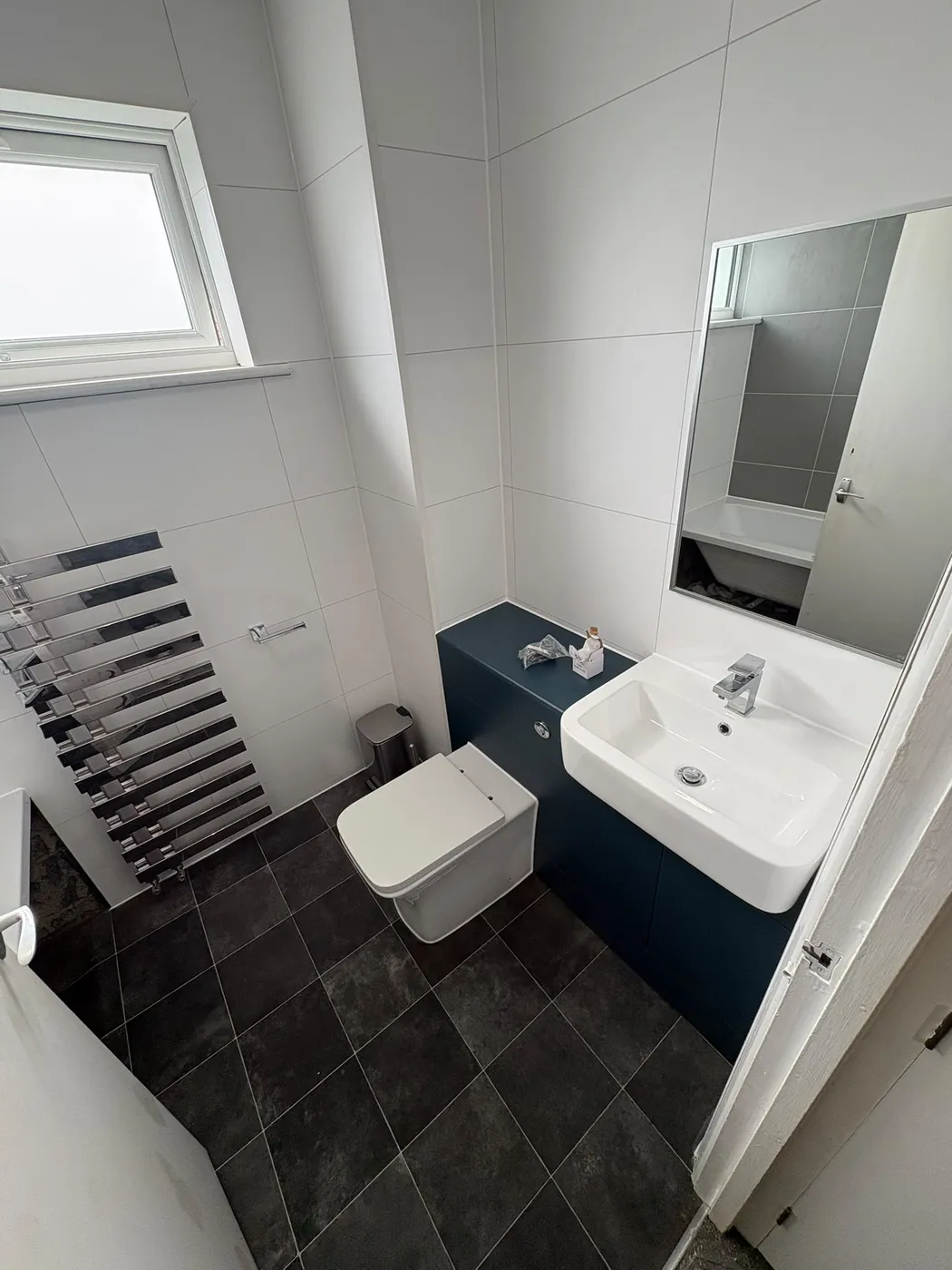 Compact modern bathroom with navy vanity unit, square basin, black towel radiator, mirrored cabinet and dark slate-effect floor tiles.