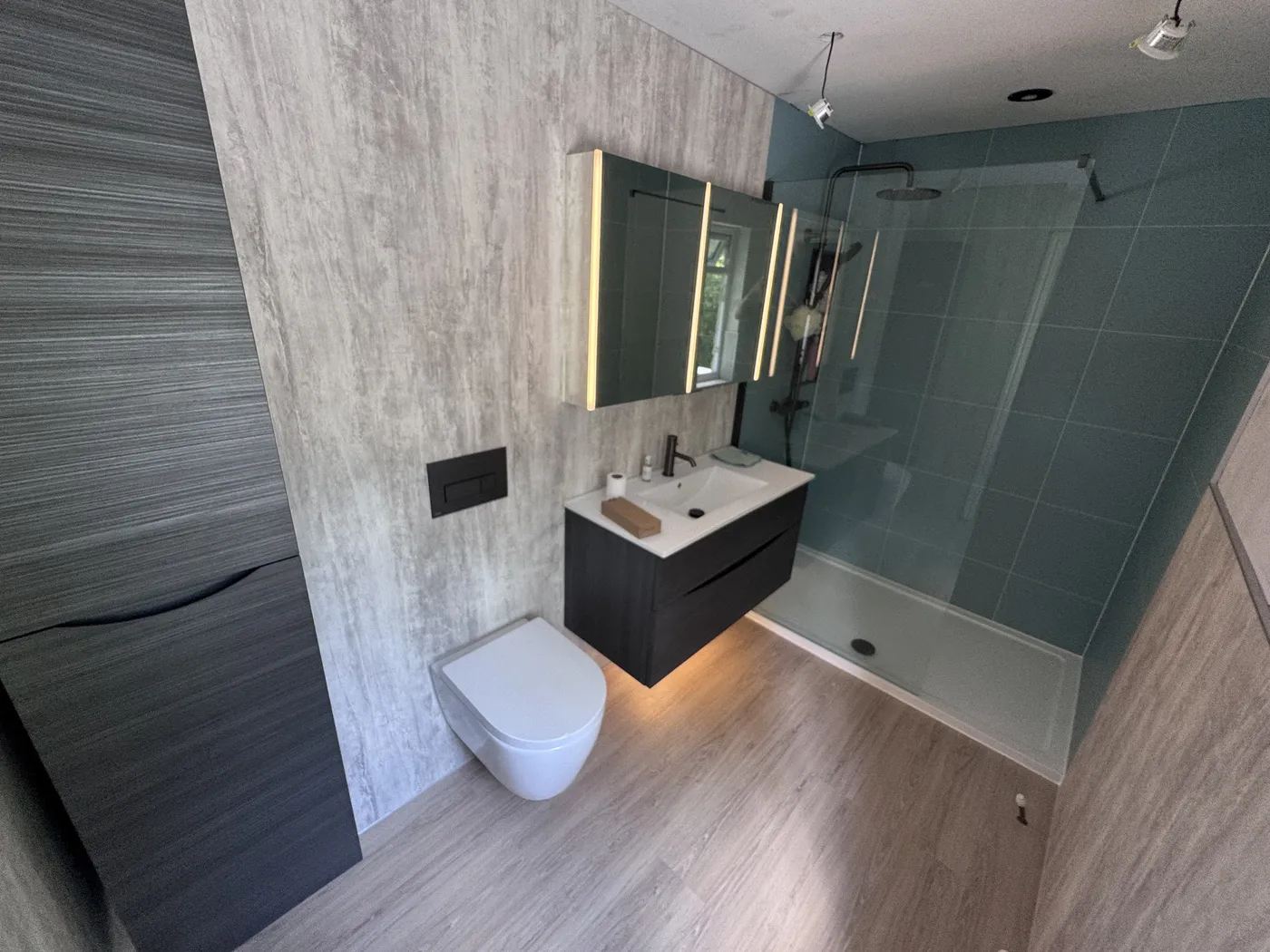 Completed bathroom renovation in London with wood-effect porcelain tiles, teal glass shower screen, wall-hung WC and backlit mirror over a floating vanity.
