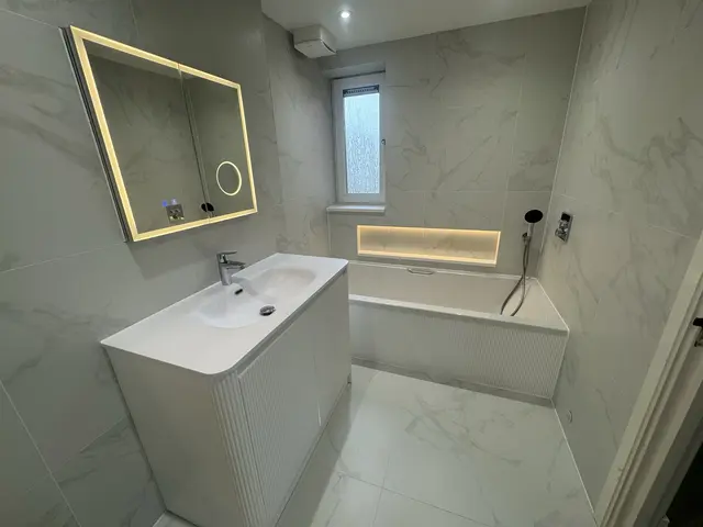 Marble-effect bathroom renovation with illuminated mirror, curved white vanity, fitted bath and LED-lit recessed niche.