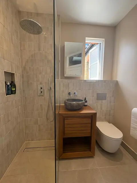 Walk-in shower with rainfall head, tiled niche, teak vanity with stone countertop basin and wall-hung WC in a beige-tiled family bathroom.