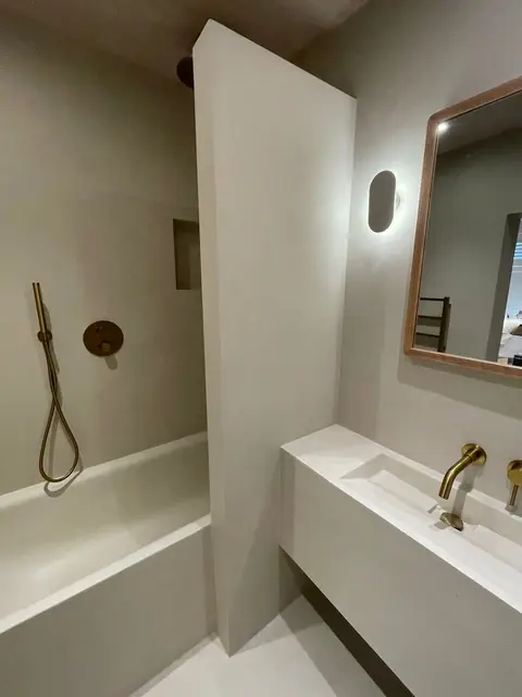 Seamless microcement bathroom in warm taupe with brushed brass shower fittings, integrated basin ledge and wood-framed mirror.