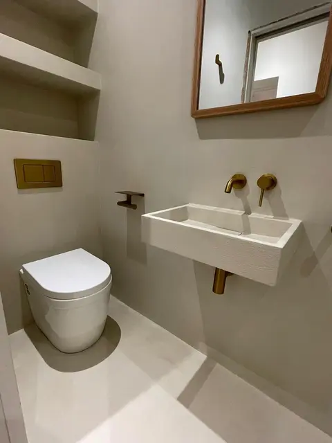 Microcement cloakroom with wall-hung WC, floating trough basin, brass wall-mounted tap and recessed shelving niches.