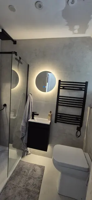 Microcement-finish shower room with matt black towel radiator, round backlit mirror, compact vanity basin and frameless glass screen.