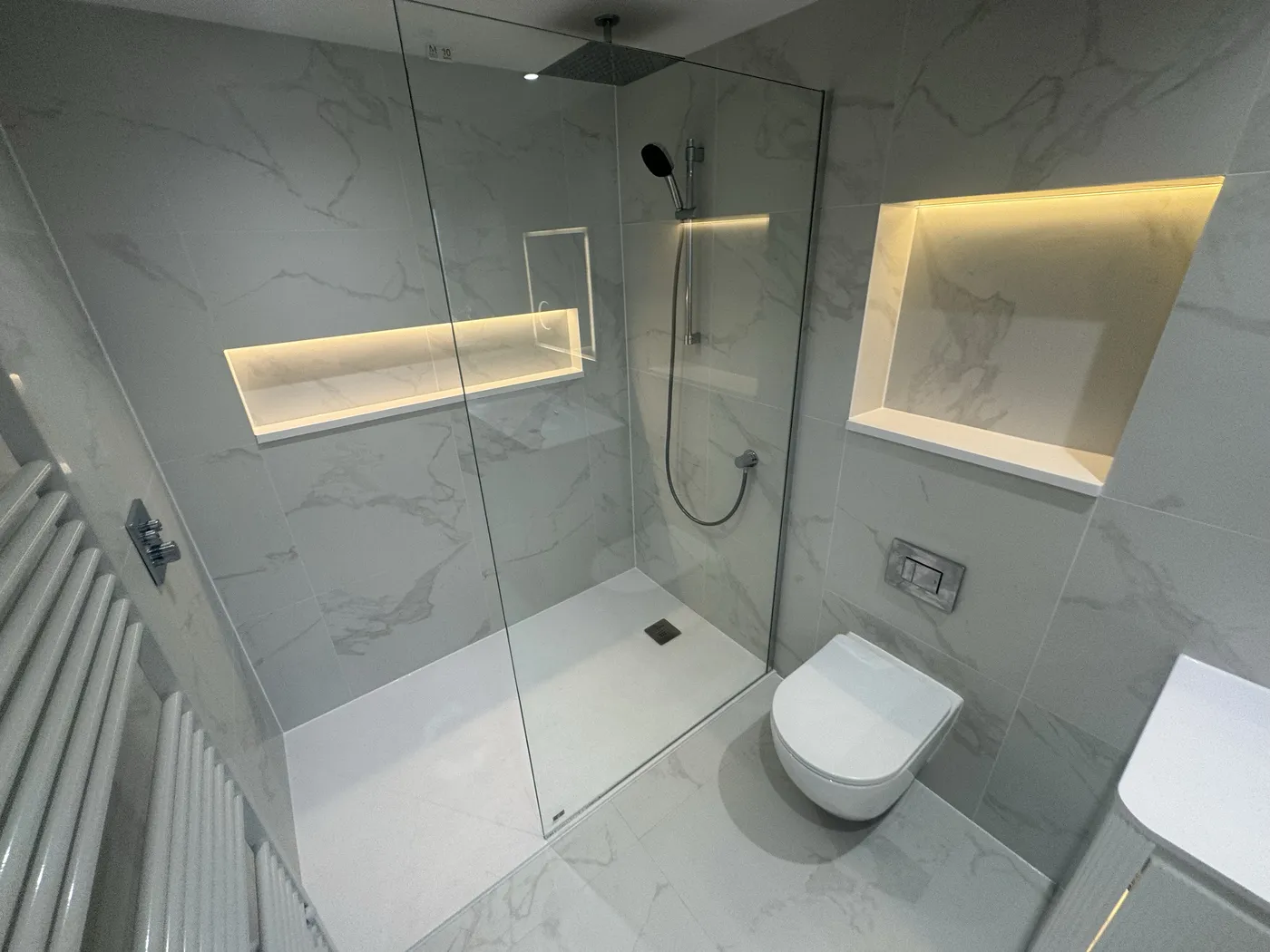 Walk-in rainfall shower in large-format marble-effect tiles with a full-width LED-lit niche and frameless glass screen.