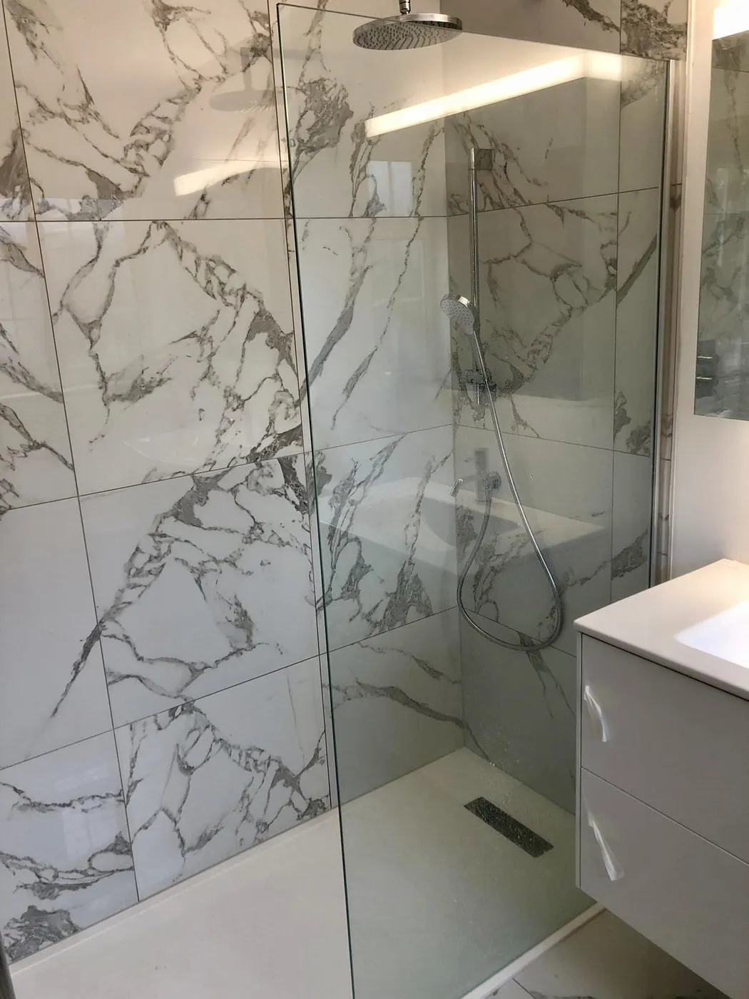 Walk-in shower enclosure in book-matched marble-effect porcelain with frameless glass panel and white floating vanity.